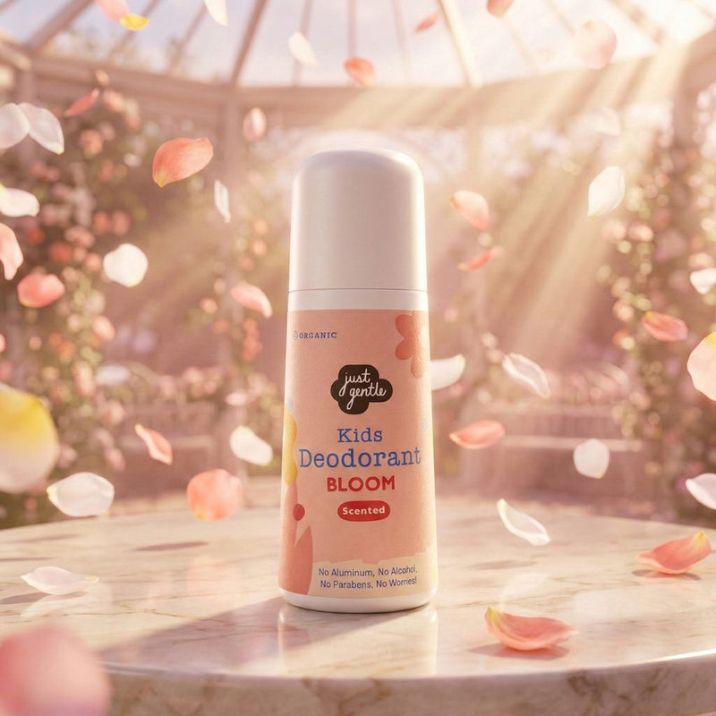 A dreamy, beautifully lit product shot of the Just Gentle 'Bloom' deodorant resting on a marble surface inside a sunlit greenhouse. Soft pink and white flower petals are floating in the air around the bottle.