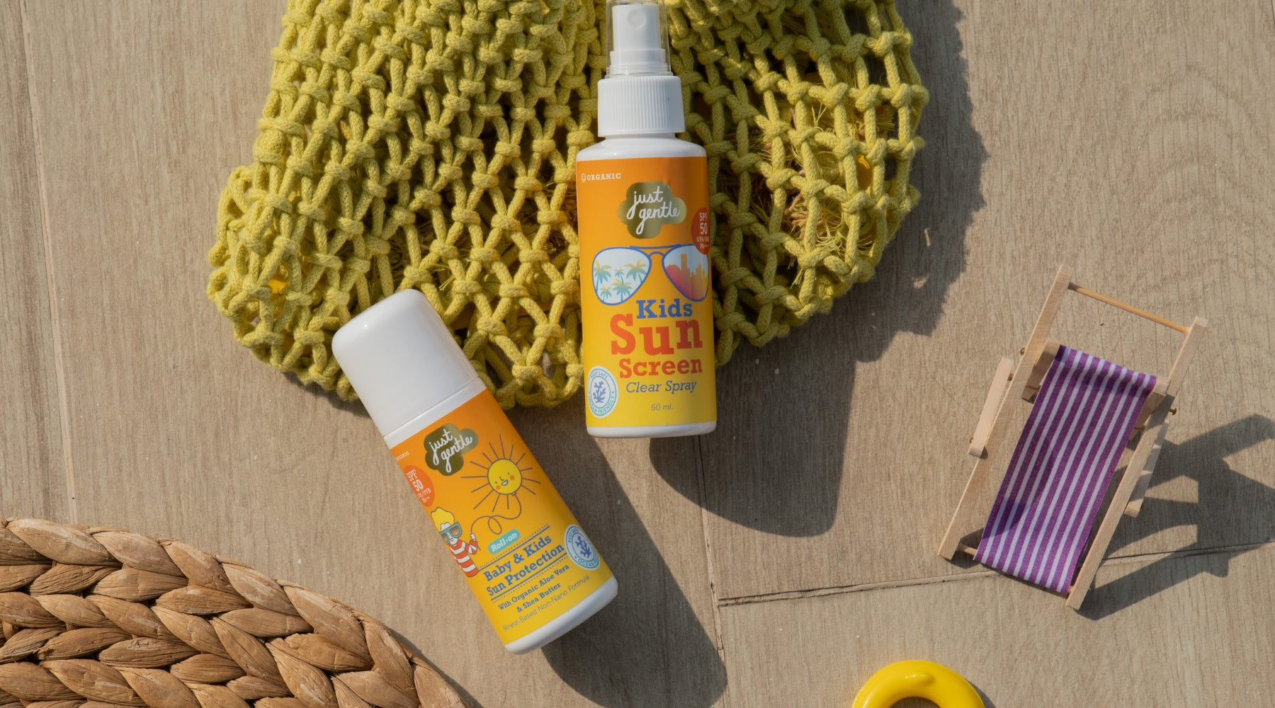 Two bottles of Just Gentle Kids' and Baby Sunscreen on a wooden surface with a yellow mesh bag and small wooden chair.