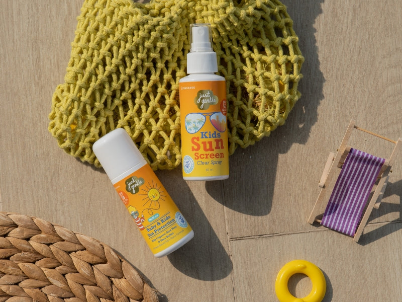 Two bottles of 'Just Gentle' kids' sunscreen with a yellow mesh bag and purple towel on a wooden surface.
