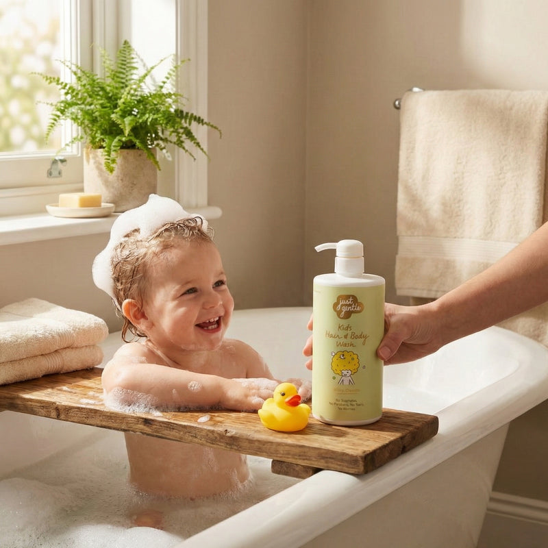 Child in a bathtub with a wooden step, holding a bottle of Just Gentle Kids Wash, with a rubber duck and towels in the background.