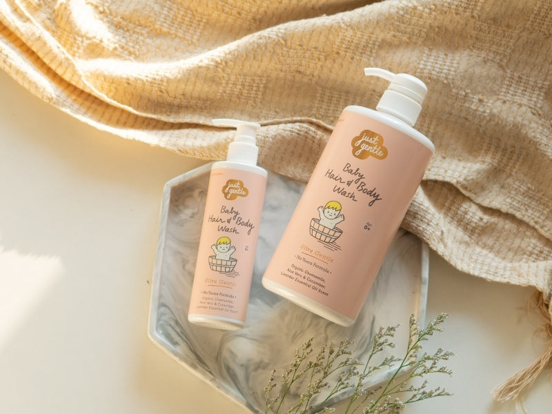 Two bottles of 'Just Gentle' baby hair and body wash on a textured surface with a beige towel and greenery. Mobile View